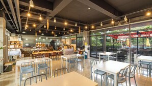 Un restaurante con mesas dentro, una zona de cocina y amplias ventanas que dan al exterior.