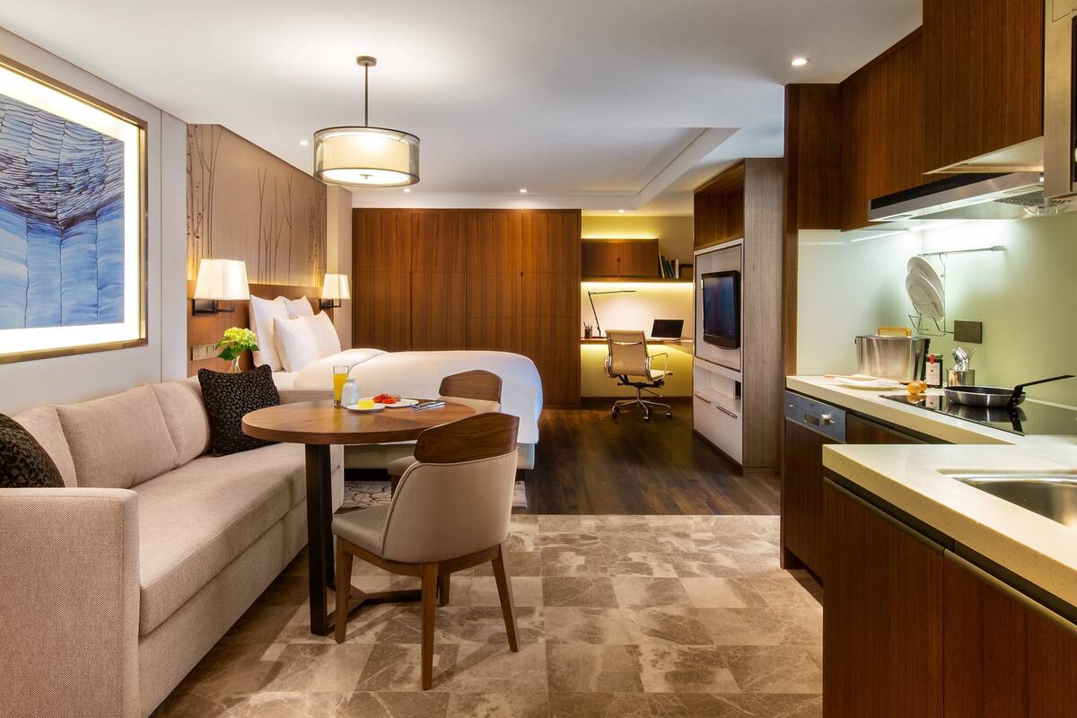 A modern hotel room with a bed, a sofa, a dining table, and a kitchen area.