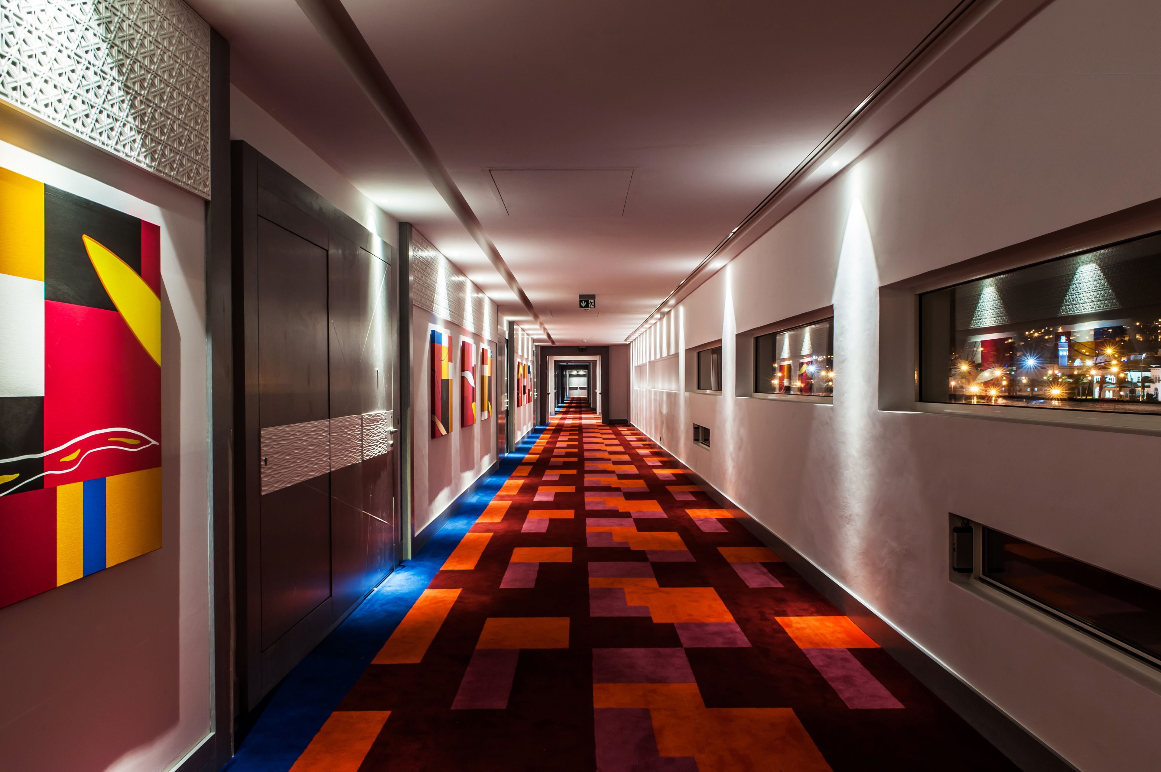 A hotel corridor with a colorful patterned carpet, modern wall art, and a view of the city at night through the windows.