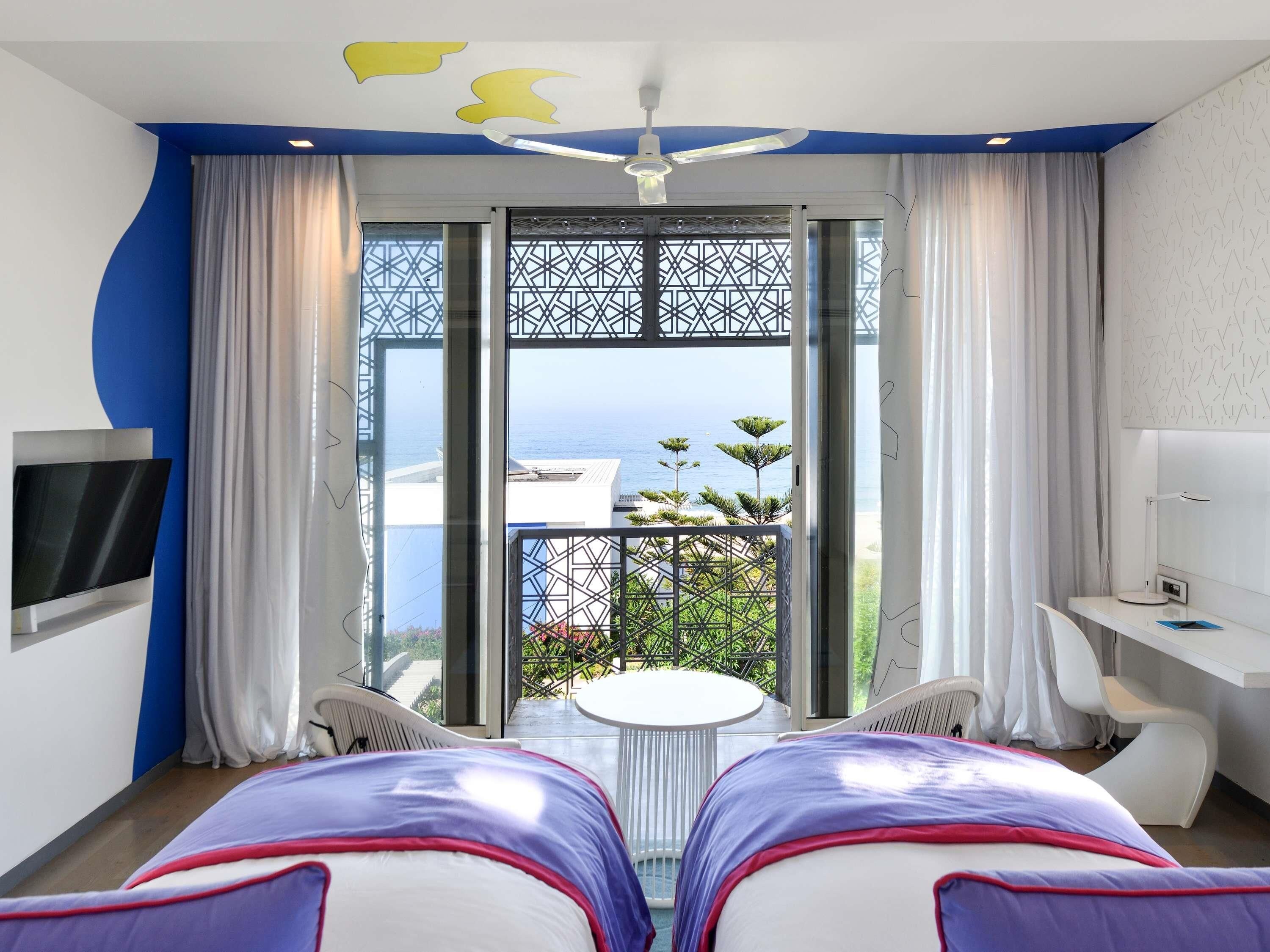 A hotel room with a bed, a desk, a TV, and a balcony with a view of the sea.