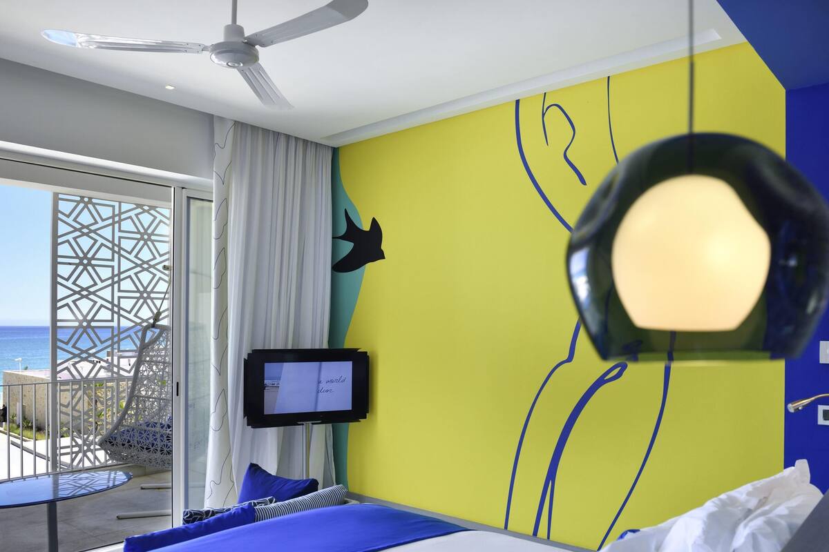 A hotel room with a bed, a TV, a ceiling fan, and a view of the ocean.
