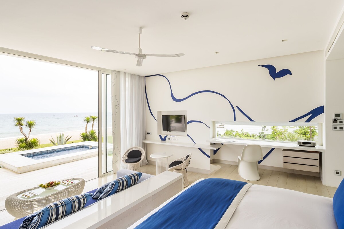 A modern hotel room with a large bed, a desk, a TV, and a view of the beach.