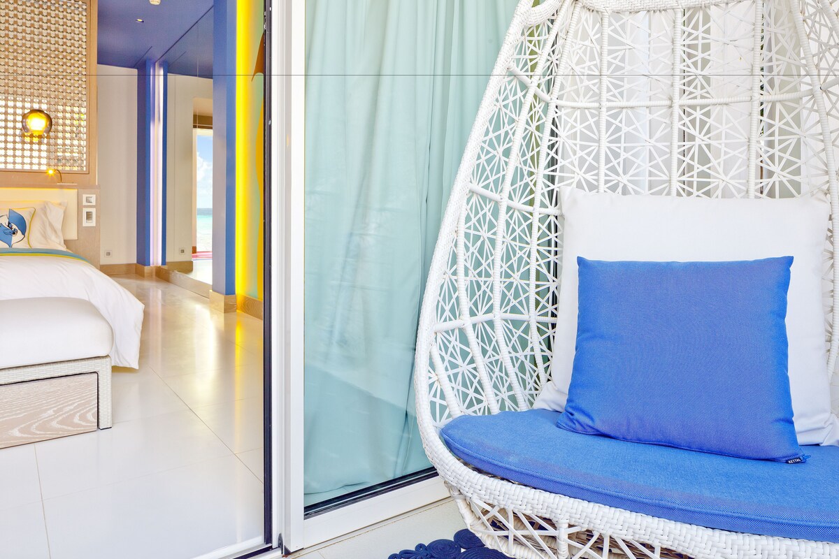 A room with a bed, a wicker chair with a blue cushion, and a glass door leading to a balcony.