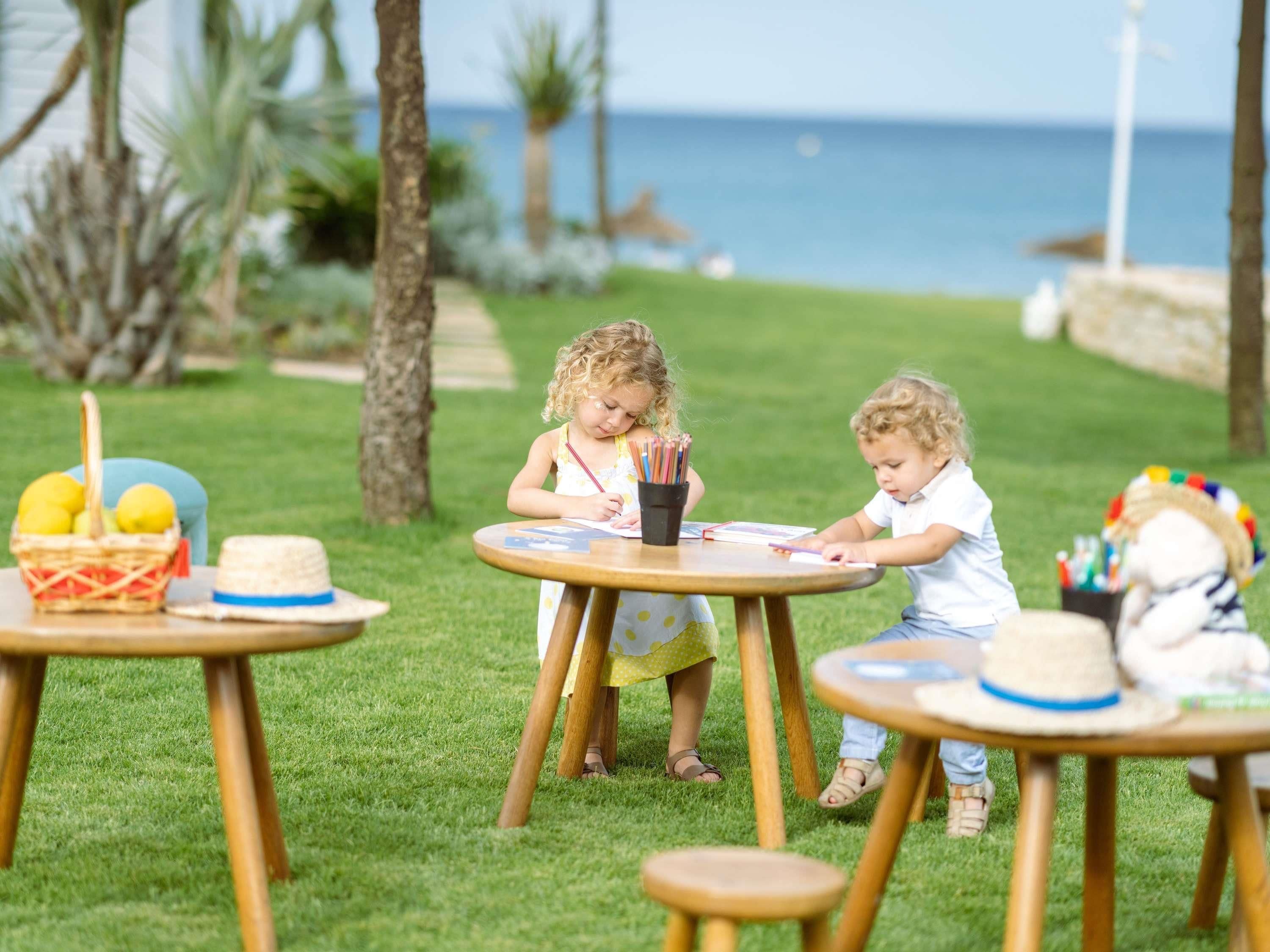 Two children at an outdoor table with art supplies, surrounded by grass and palm trees, with the ocean in the background.
