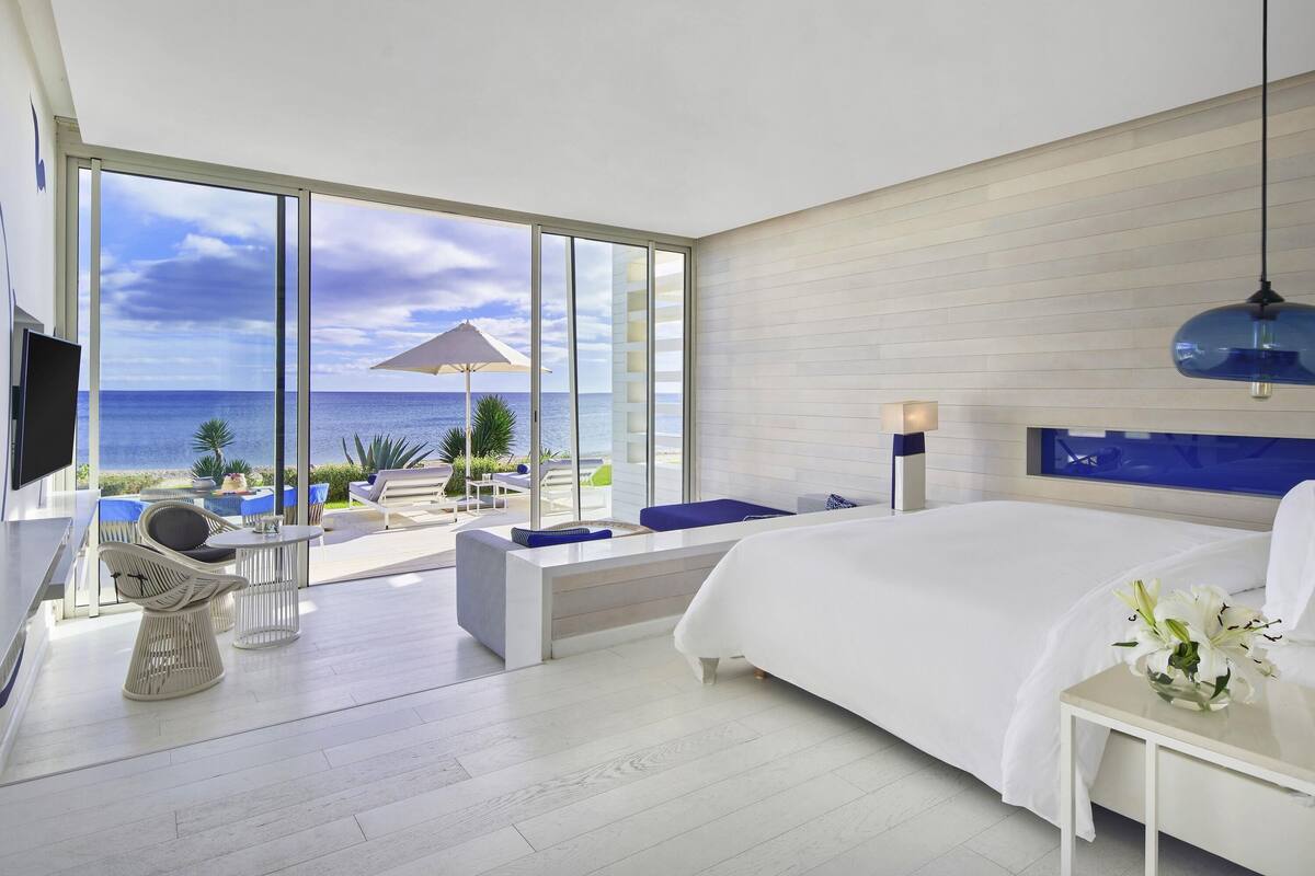 A modern hotel room with a large bed, a TV, a balcony with a view of the sea, and a dining area with chairs and a table.