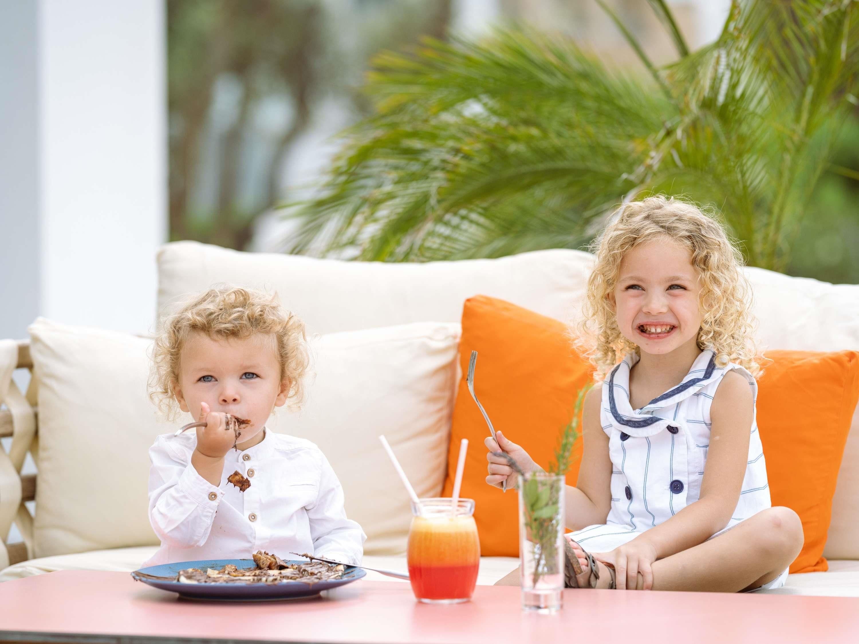 Two children sitting on a white sofa with orange cushions, a table with a plate of food and a drink, and a glass of water with a straw.