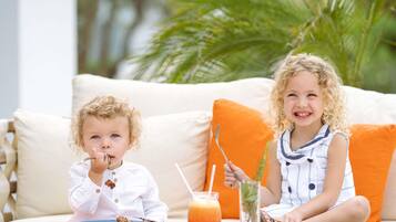 Two children sitting on a white sofa with orange cushions, a table with a plate of food and a drink, and a glass of water with a straw.