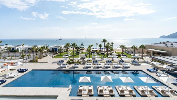Un área de piscina con sillones de descanso y sombrillas, vista al mar y un edificio con un mural.
