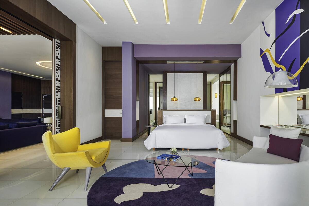 A modern hotel room with a bed, a yellow armchair, a white sofa, a glass coffee table, and a purple accent wall.