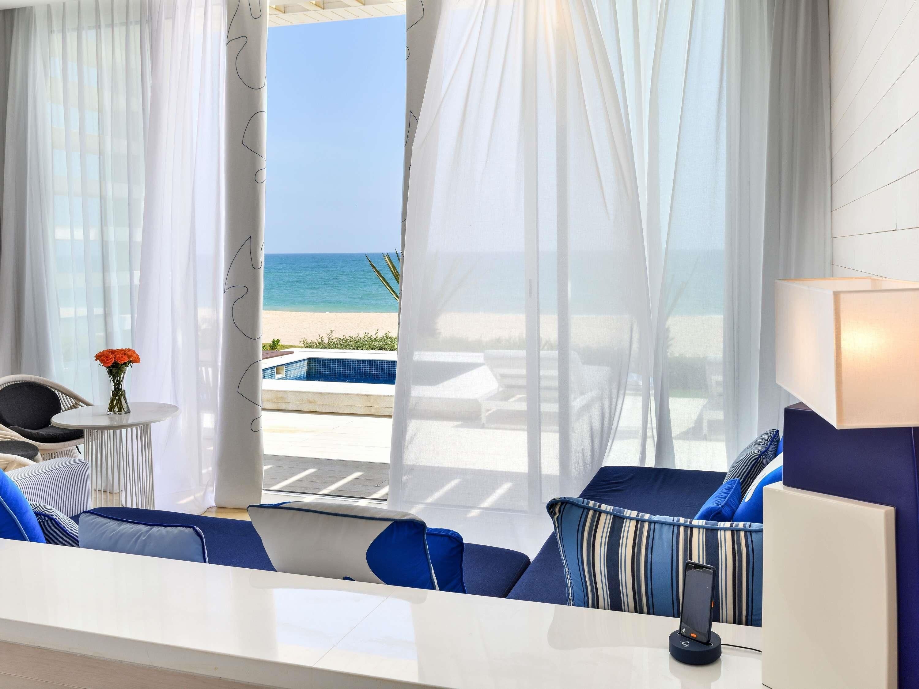 Ein modernes Hotelzimmer mit Blick auf den Strand, einen Pool und eine Essecke mit Tisch und Stühlen.