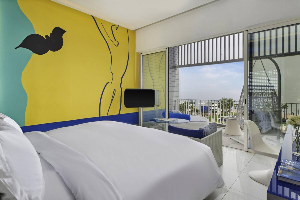 A hotel room with a bed, a TV, a balcony with seating, and a view of the city.