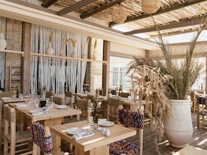 An outdoor dining area with wooden tables and chairs, a thatched roof, and decorative plants.