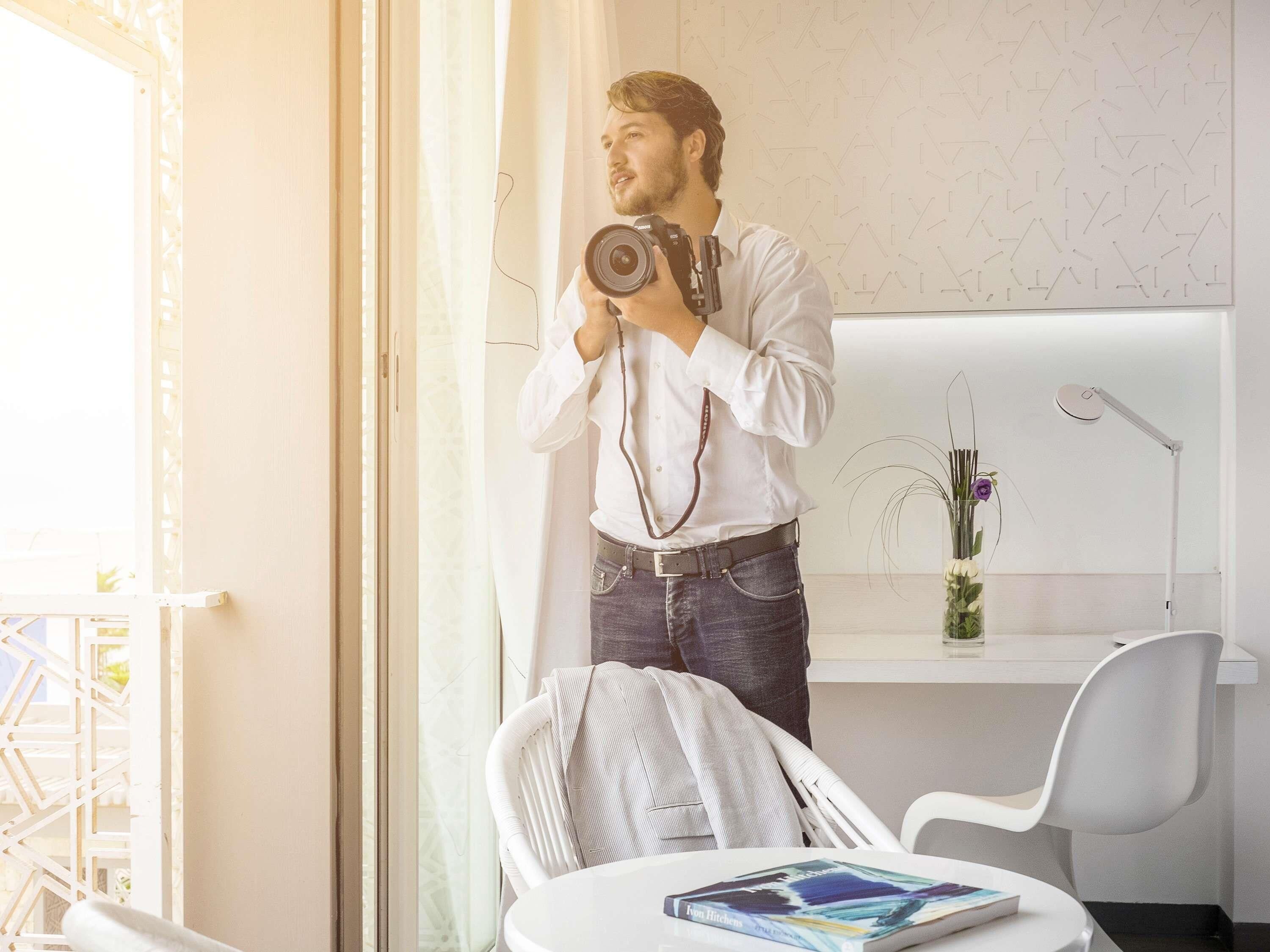 A person in a white shirt and jeans holding a camera in a room with a white chair, a white desk, and a window with a view.