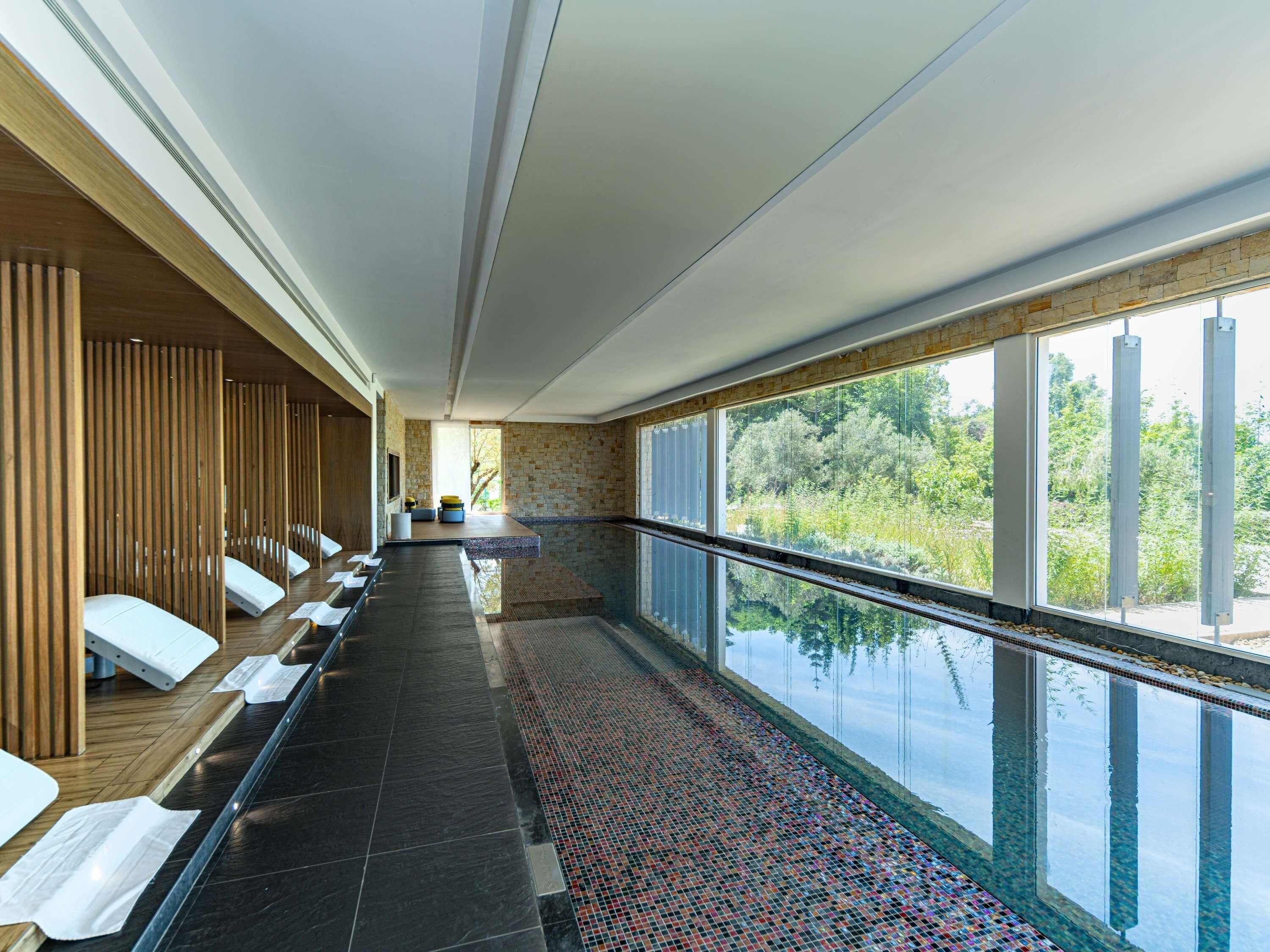 Une longue et étroite piscine intérieure, avec un sol carrelé, des bancs en bois d’un côté et de grandes fenêtres de l’autre.
