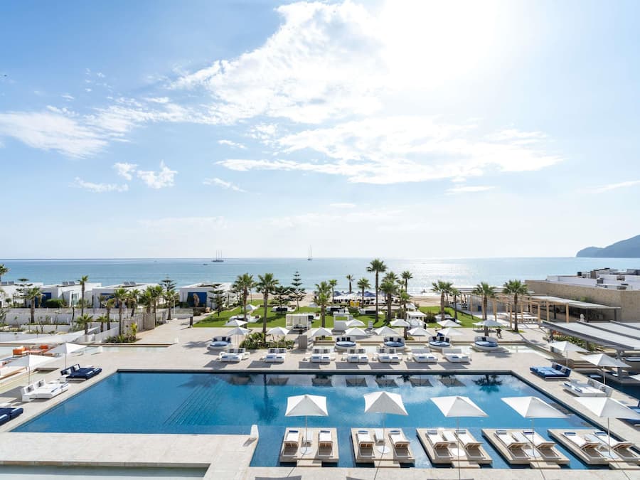 A pool area with lounge chairs and umbrellas, a view of the sea, and a building with a mural.