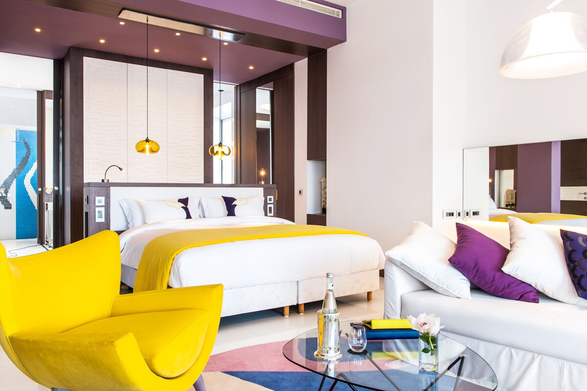 A modern hotel room with a large bed, a sofa, a glass coffee table, and a yellow armchair.