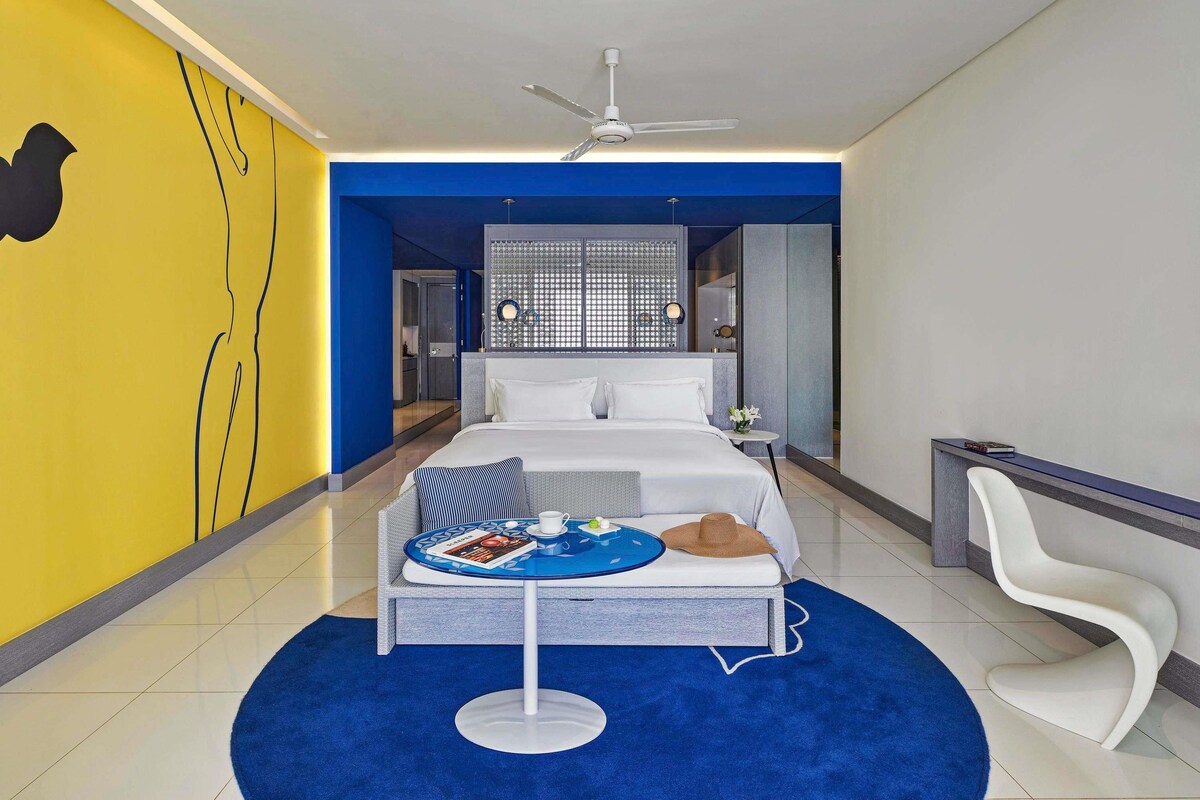 A modern hotel room with a bed, a desk, and a round table with a book and a cup.