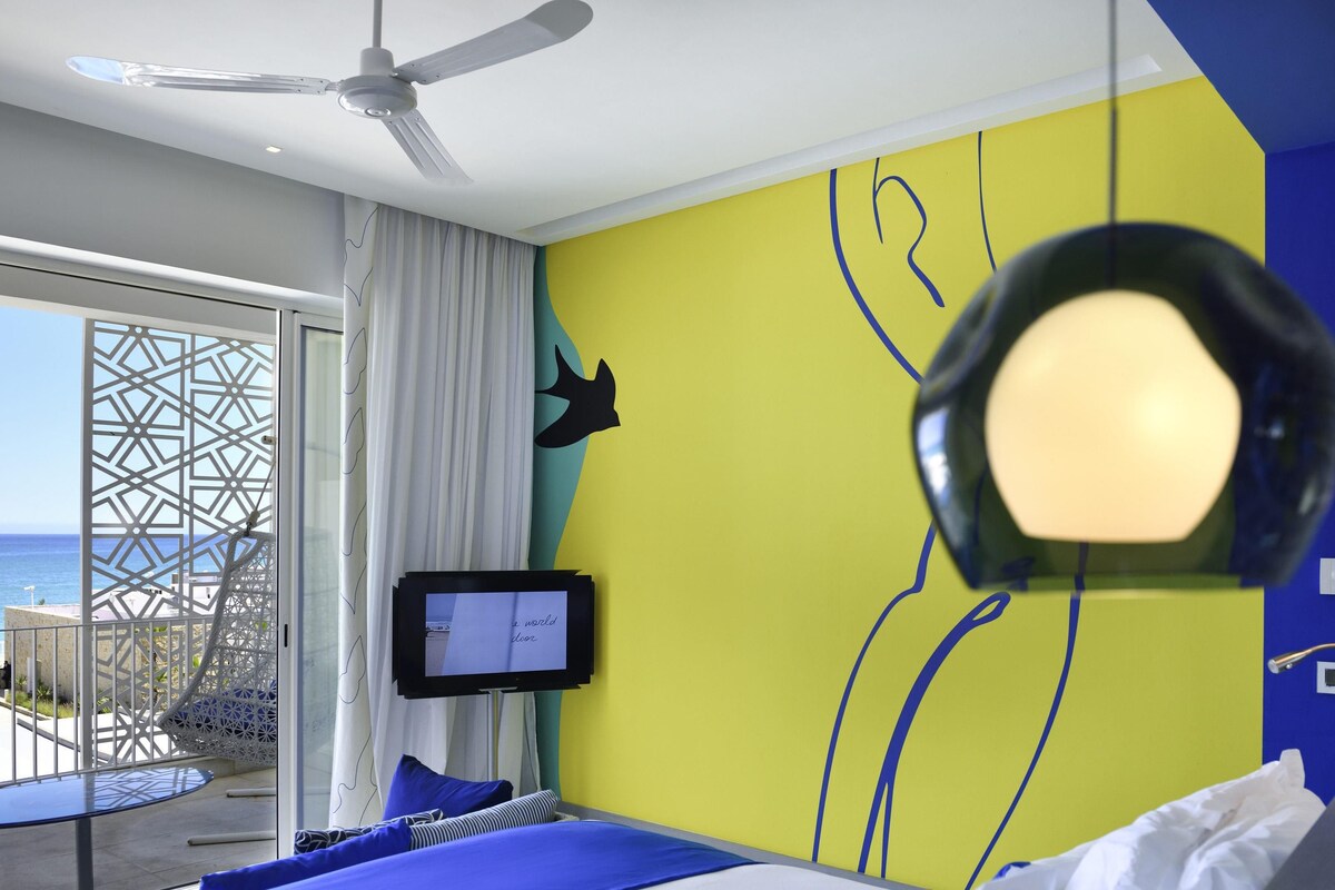 Ett hotellrum med en säng, en TV, en takfläkt och utsikt över havet.