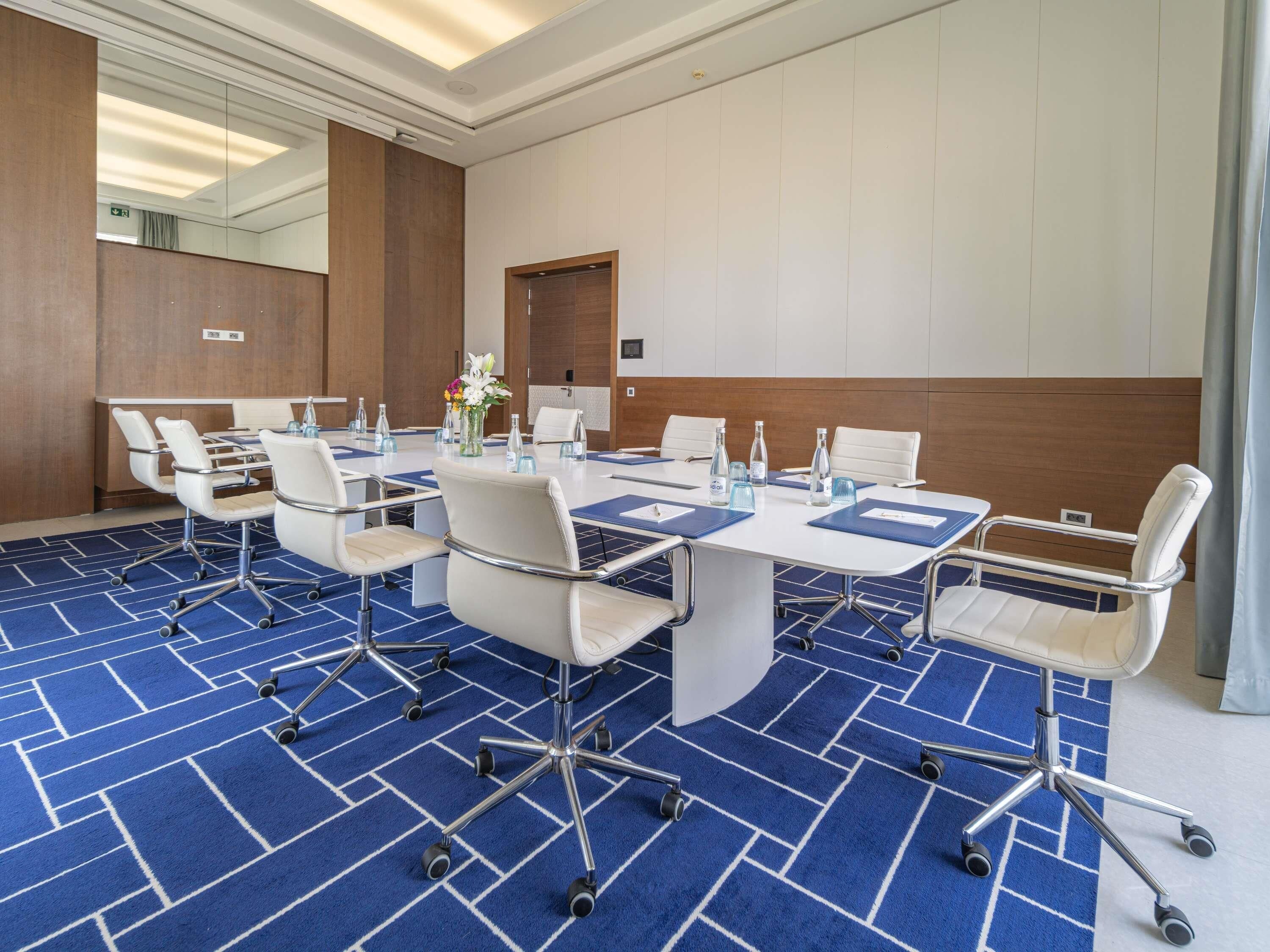 Une salle de conférence avec une grande table, des chaises blanches et une moquette bleue.