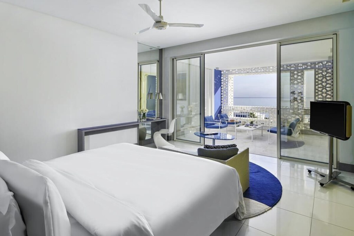 A hotel room with a bed, a TV, a ceiling fan, a large window with a view of the city, and a balcony with seating.