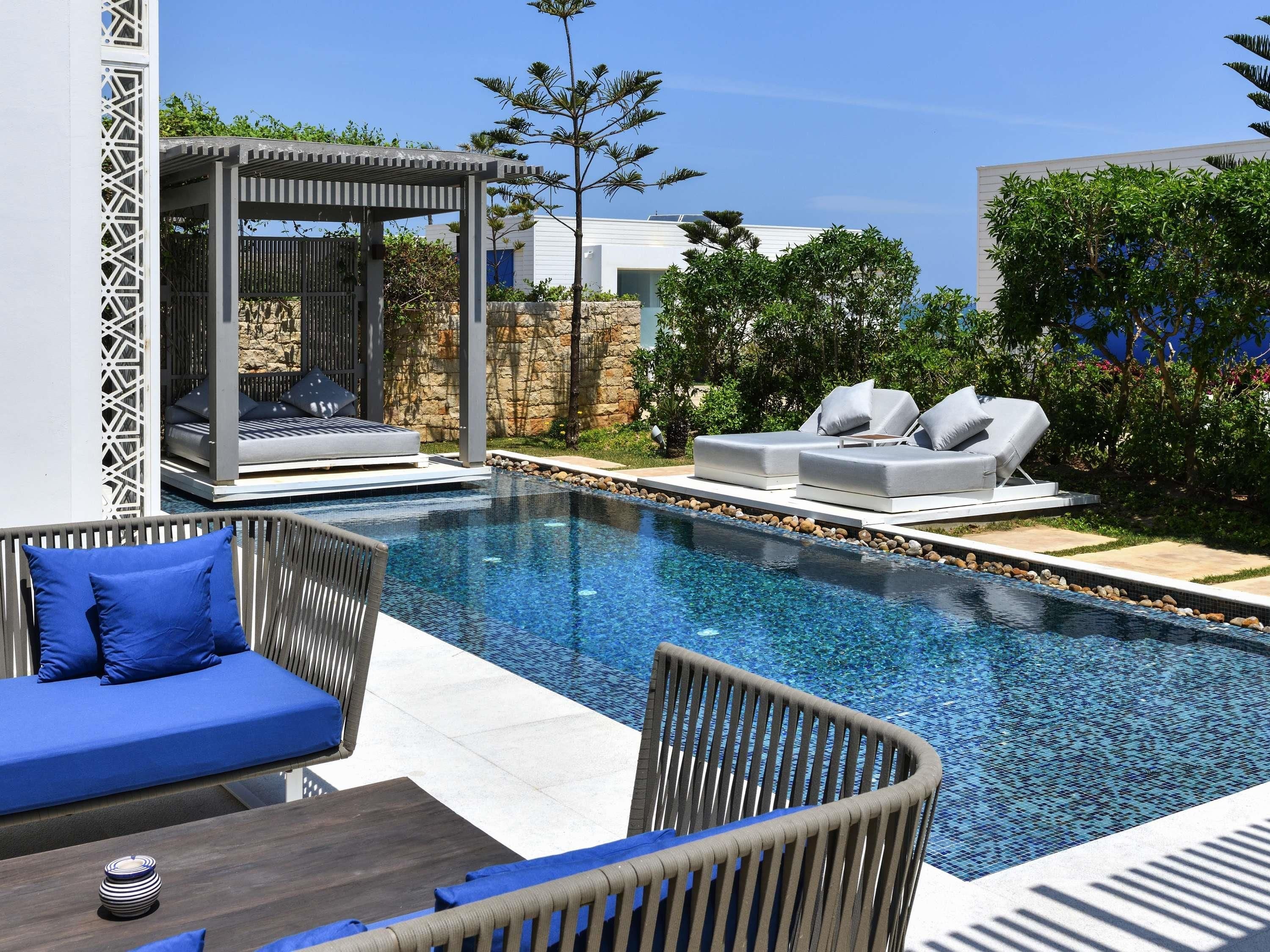 A swimming pool with lounge chairs, a covered seating area, and a stone wall.