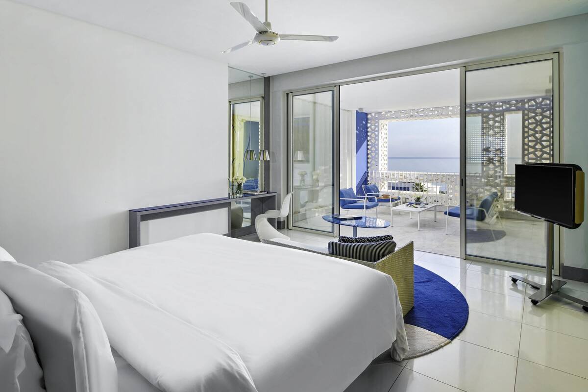 A hotel room with a bed, a TV, a balcony with seating, and a view of the sea.