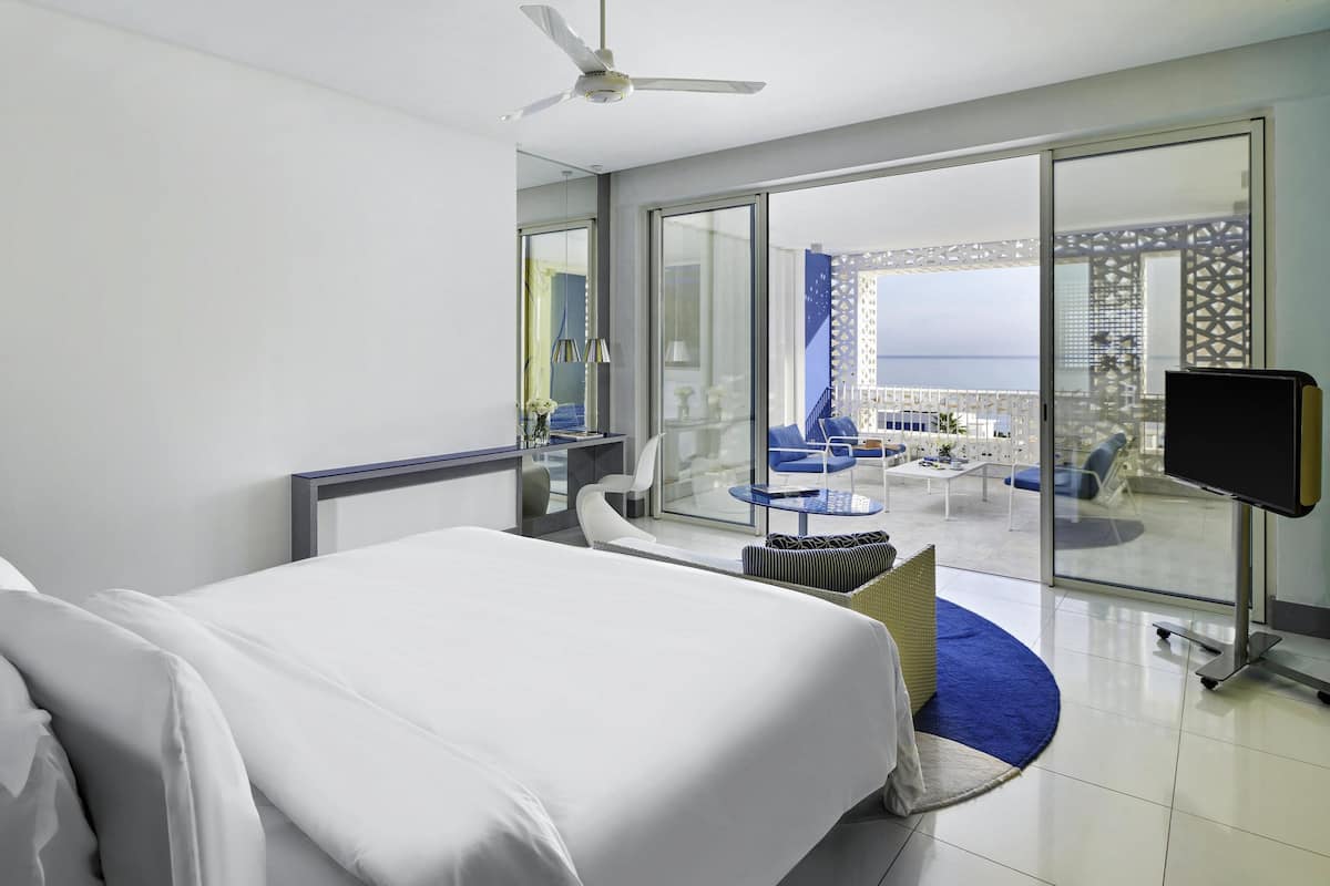 A hotel room with a bed, a TV, a balcony with seating, and a view of the sea.