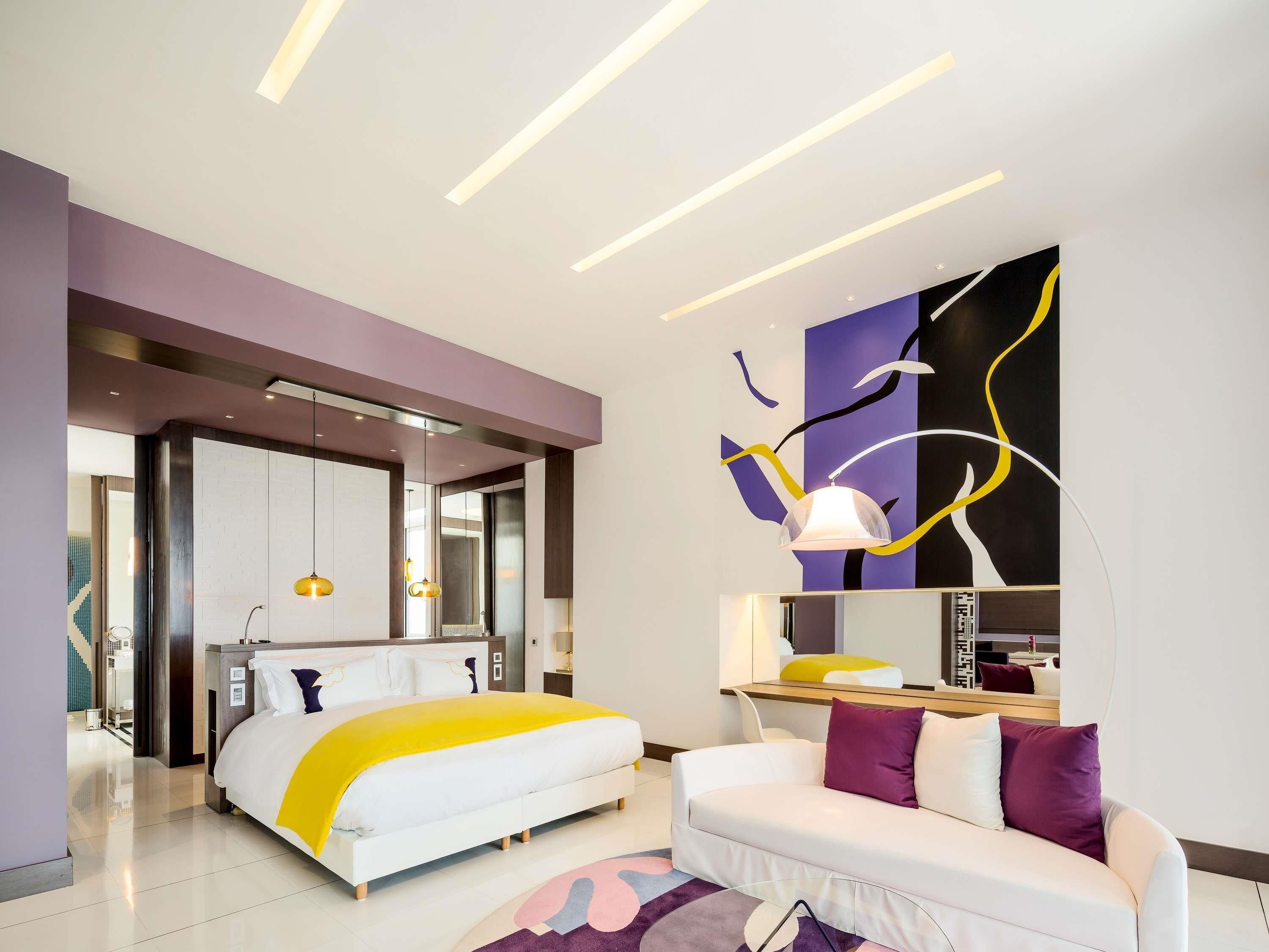 Une chambre d’hôtel moderne, équipée d’un lit, d’un canapé, d’un bureau et ornée d’un grand tableau abstrait au mur.