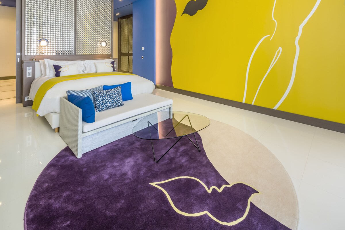 A modern hotel room with a bed, a sofa, a ceiling fan, and a large wall mural.