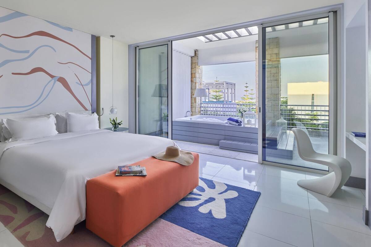 A modern hotel room with a large bed, a balcony with a view, and a seating area with a chair.