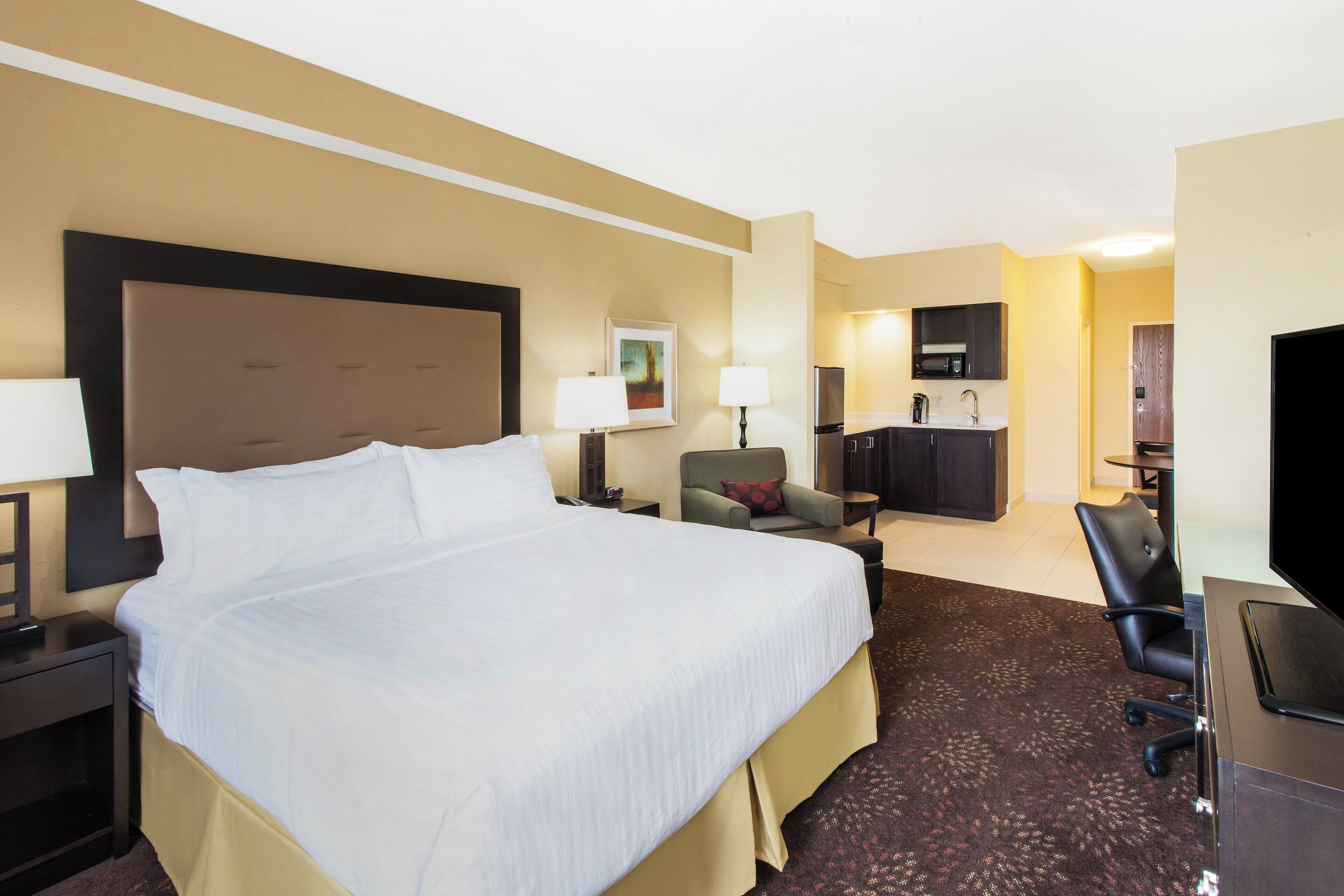 A hotel room with a large bed, a desk, a chair, a sofa, a television, and a kitchenette.