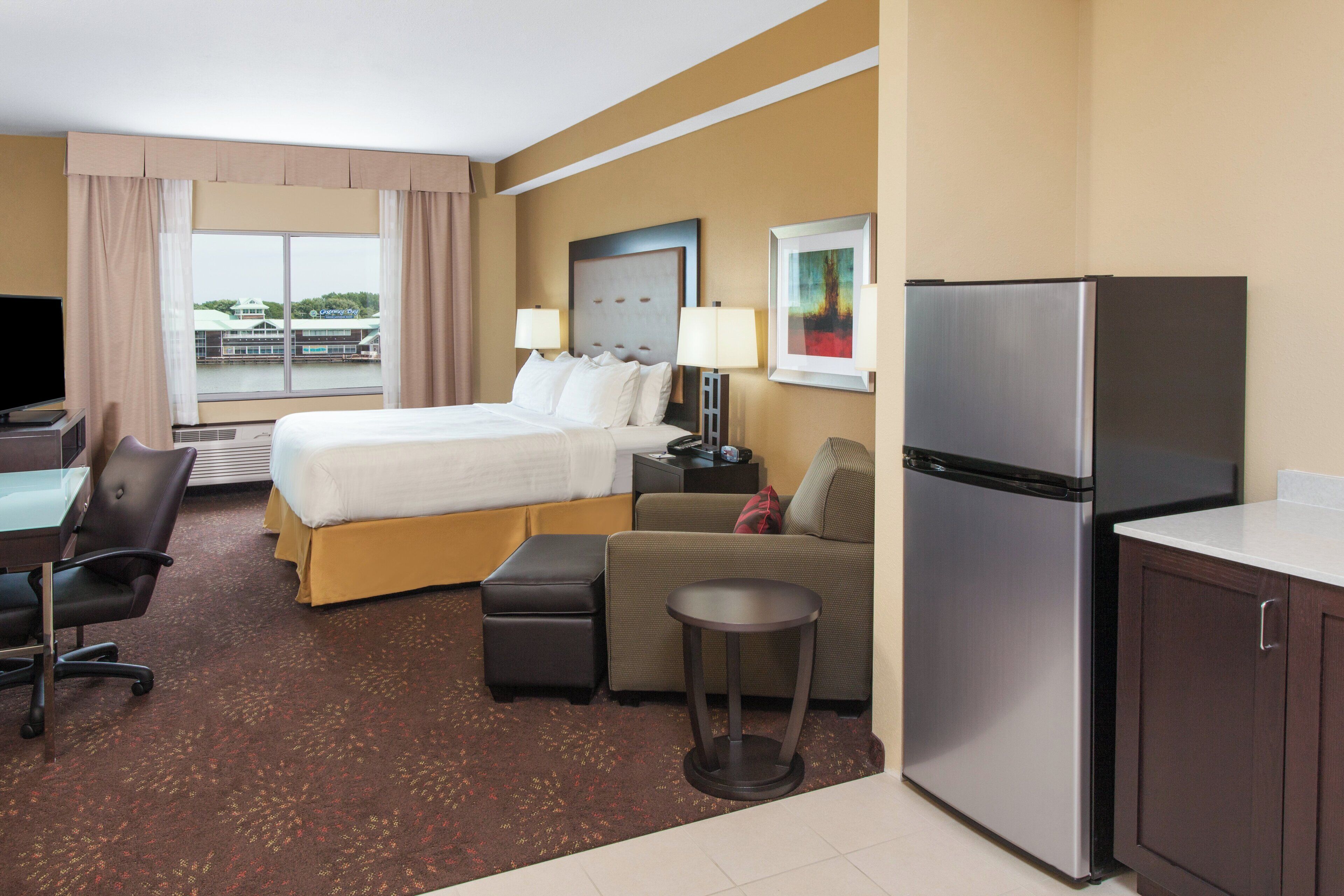 Habitación de hotel con cama, escritorio, silla, refrigerador y vista a un edificio.