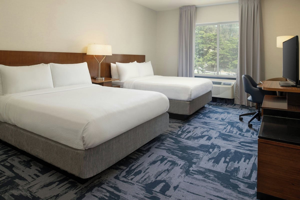 A hotel room with two beds, a desk with a computer, a lamp, and a view of trees through the window.