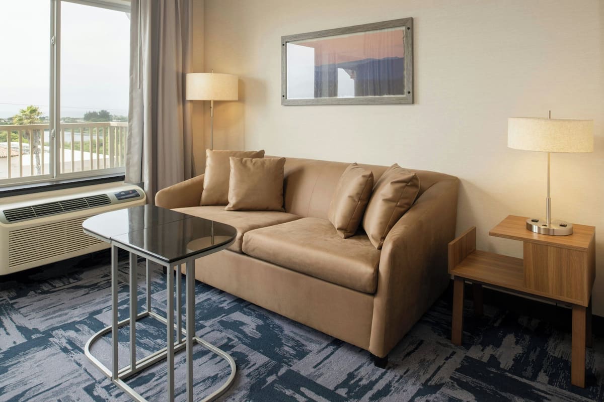 A hotel room with a beige sofa, a small round table, two bedside lamps, and a large window with a view of the outdoors.