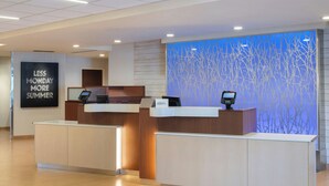 A hotel reception area with two reception desks, a digital check-in kiosk, a wall with a 'Less Monday More Summer' sign, and a blue decorative wall. - Fairfield Inn & Suites by Marriott Santa Cruz, CA (Santa Cruz)