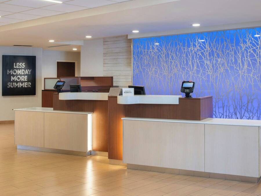 A hotel reception area with two reception desks, a digital check-in kiosk, a wall with a 'Less Monday More Summer' sign, and a blue decorative wall.