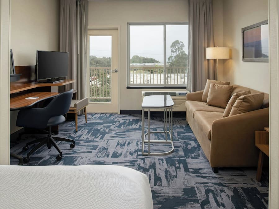 A hotel room with a bed, a desk with a computer, a couch, and a view of the outdoors through a balcony.