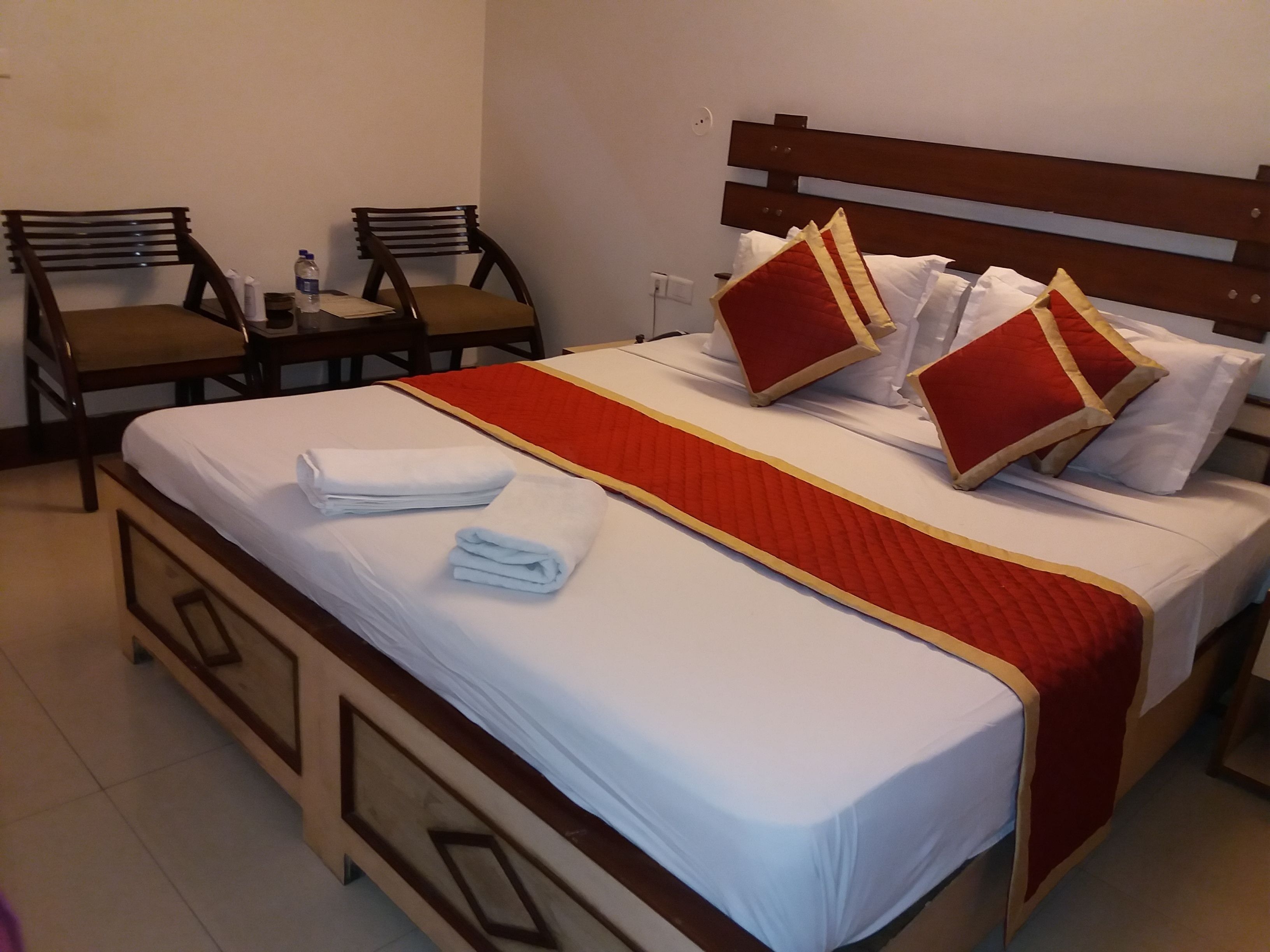 A neatly made bed with white linens and red pillows, a wooden headboard, and two chairs with brown cushions.