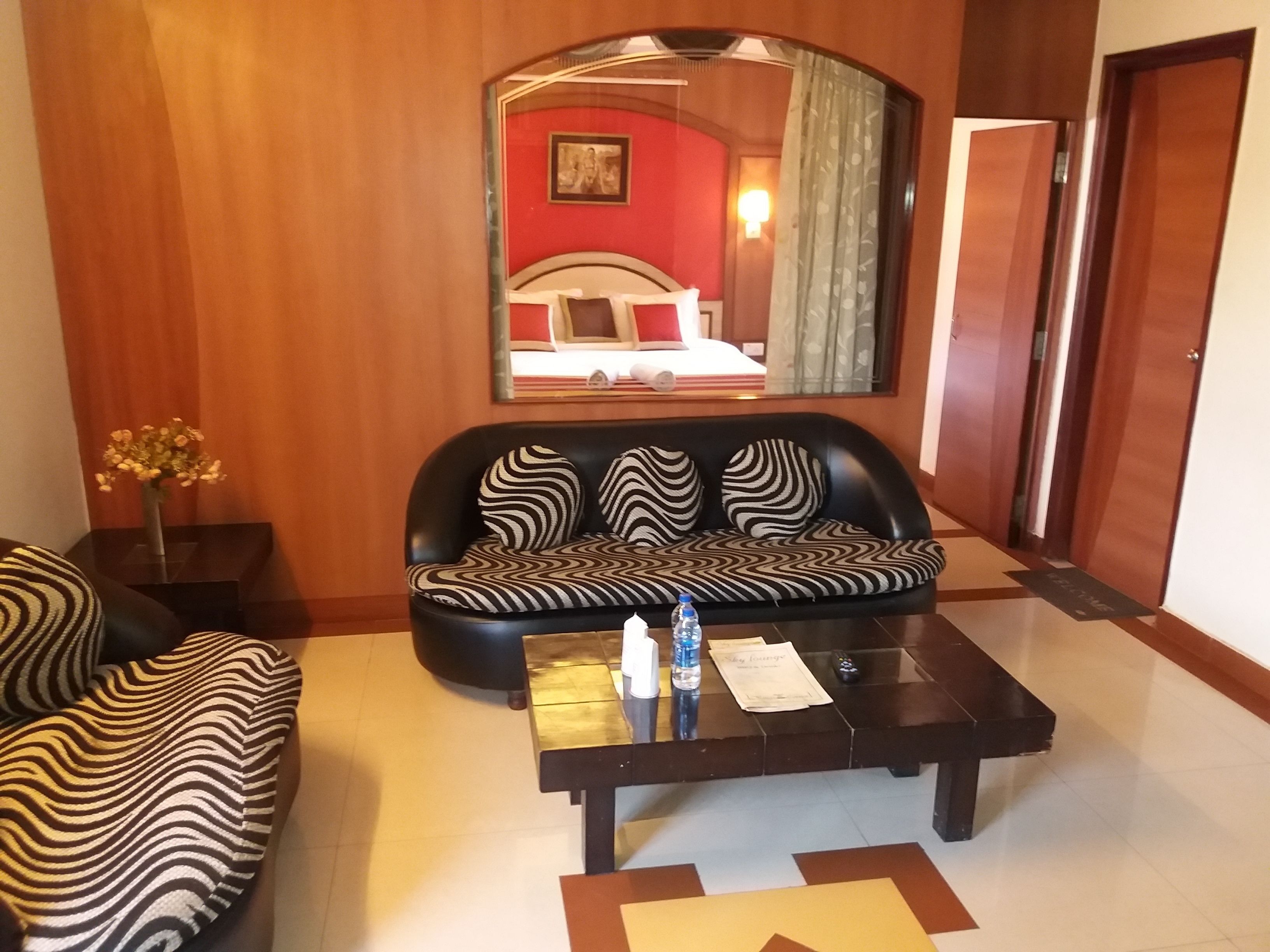 Habitación de hotel con sofá negro, una mesita de centro de madera, un espejo y una cama frente a una pared roja con un cuadro.