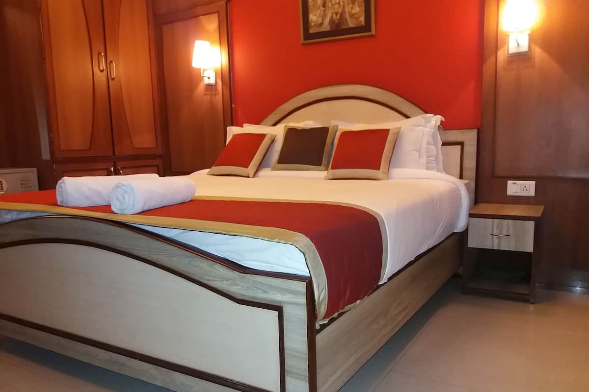 Cama bem arrumada com colcha vermelha e branca, dois travesseiros e uma toalha enrolada. Há uma cabeceira de madeira, uma mesa de cabeceira com abajur e um ponto de luz fixado na parede.