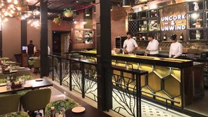 Restaurante com área de bar, mesas com cadeiras estofadas em verde, plantas pendentes e parede de tijolos.