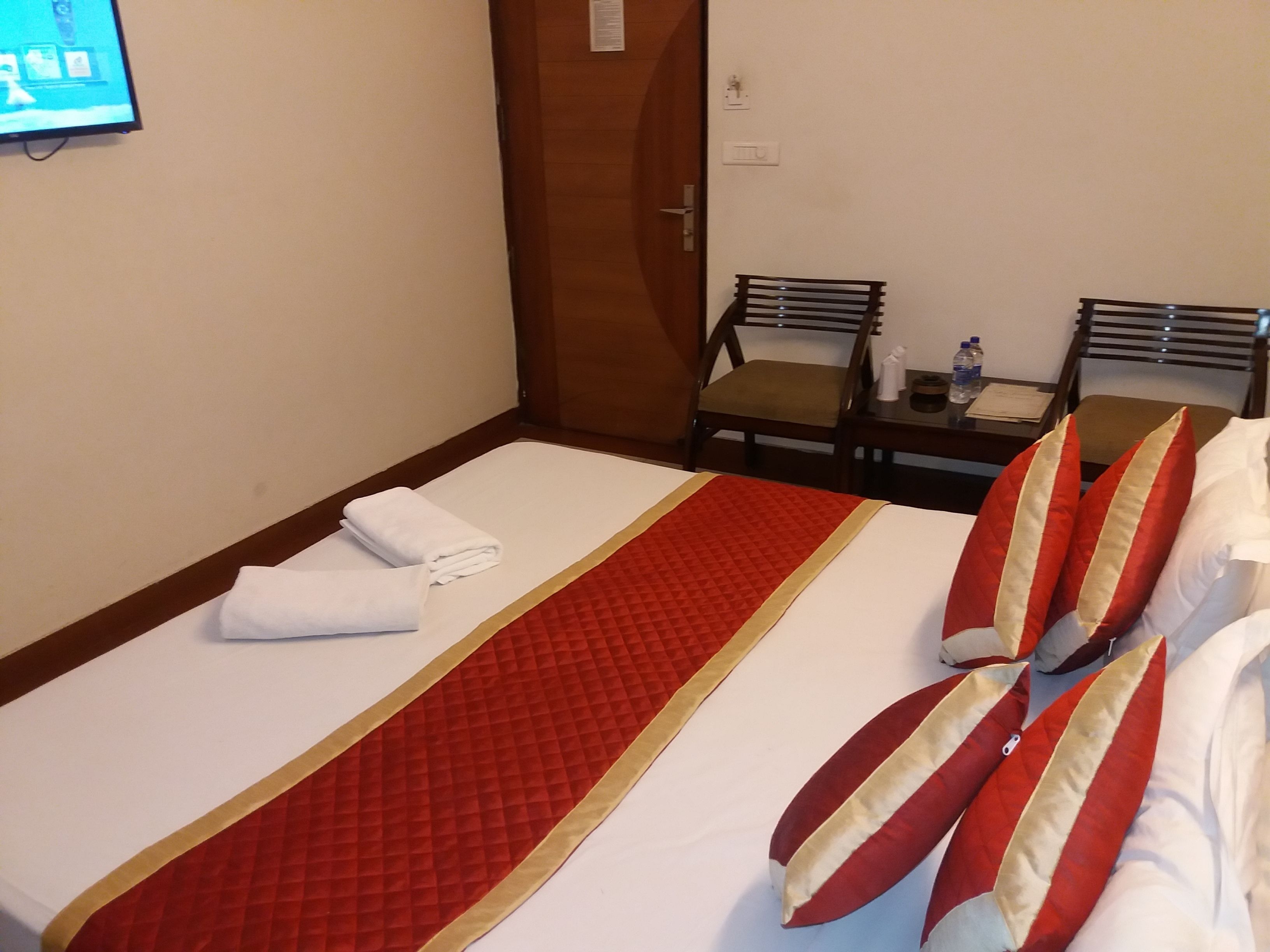 Habitación de hotel con una cama, dos sillas, una mesita con una botella y pañuelos, y un televisor en la pared.