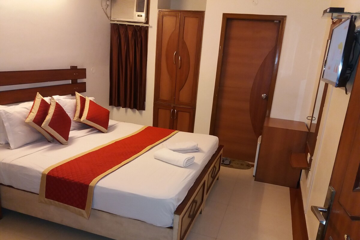 A neatly made bed with white linens and red accents, a wooden wardrobe, and a television mounted on the wall.