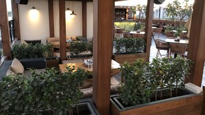 Uma área externa com mesas e cadeiras de madeira, rodeada por floreiras com vegetação.