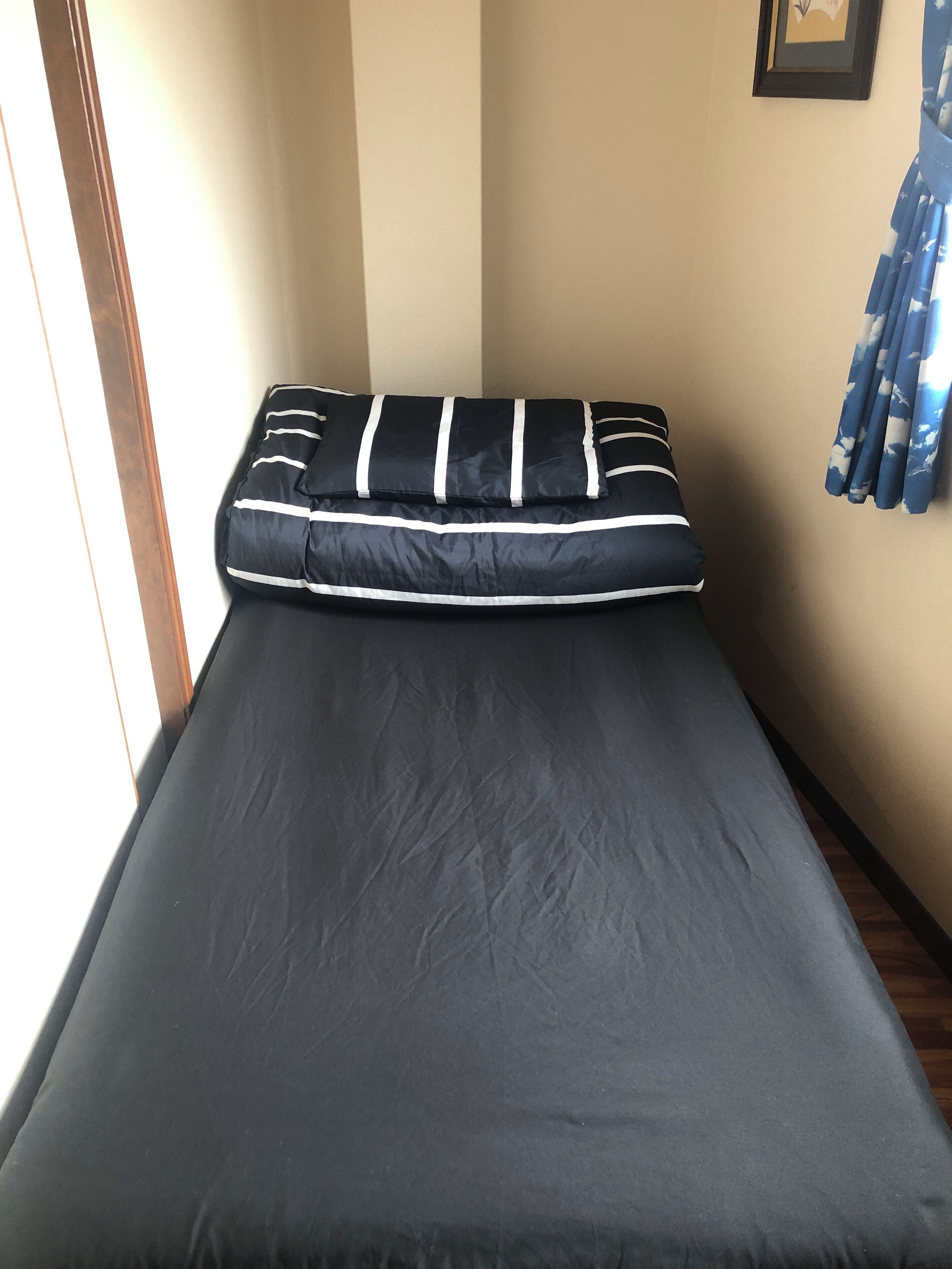 A small bed with a black mattress and white-striped pillow in a narrow room with beige walls and a window on the left.