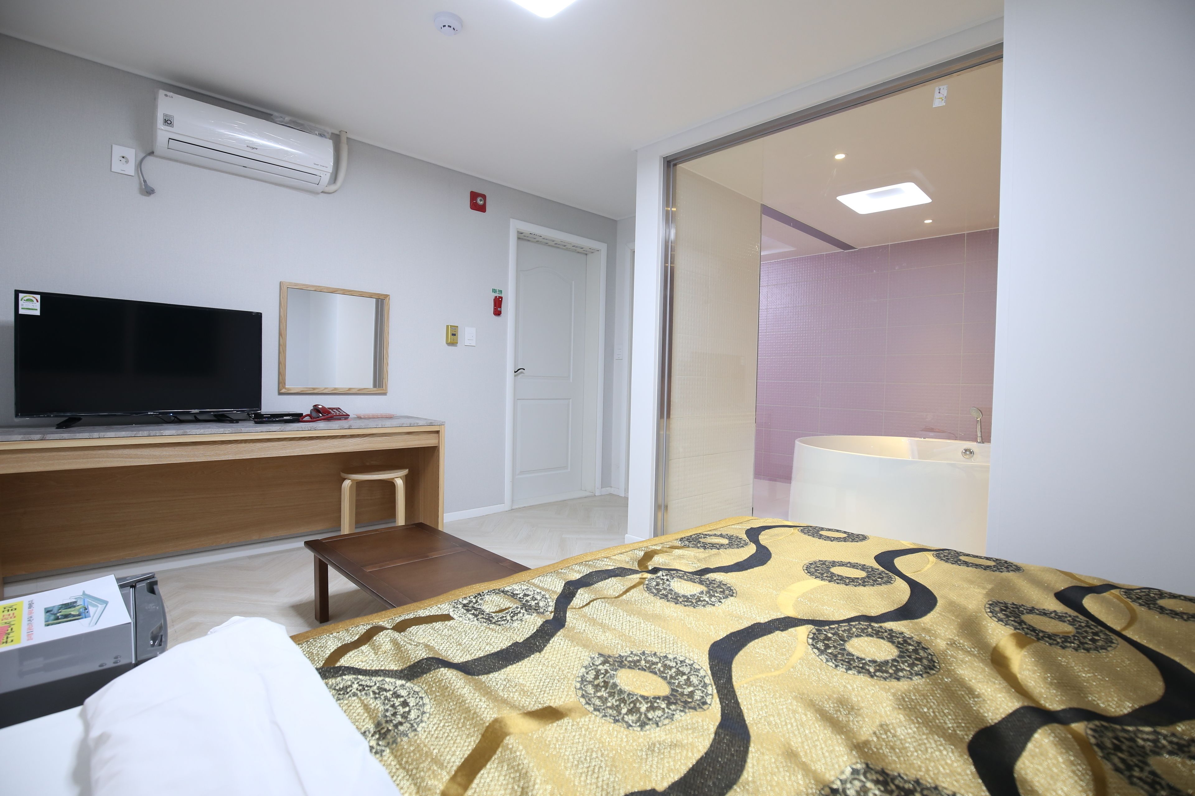 A hotel room with a bed, a desk with a flat-screen TV, a small table, an air conditioning unit, and a bathroom visible through an open door.