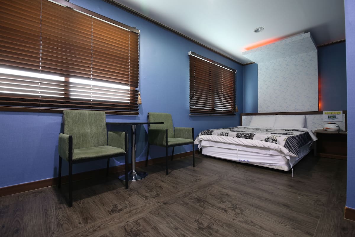 A room with a bed, two chairs, a table, and two windows with blinds.