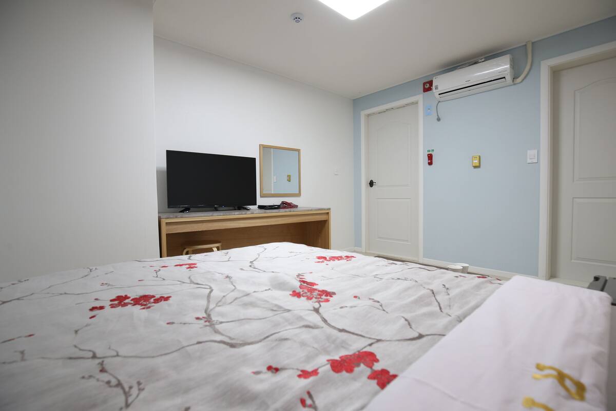 A bedroom with a bed, a desk, a television, and an air conditioning unit.