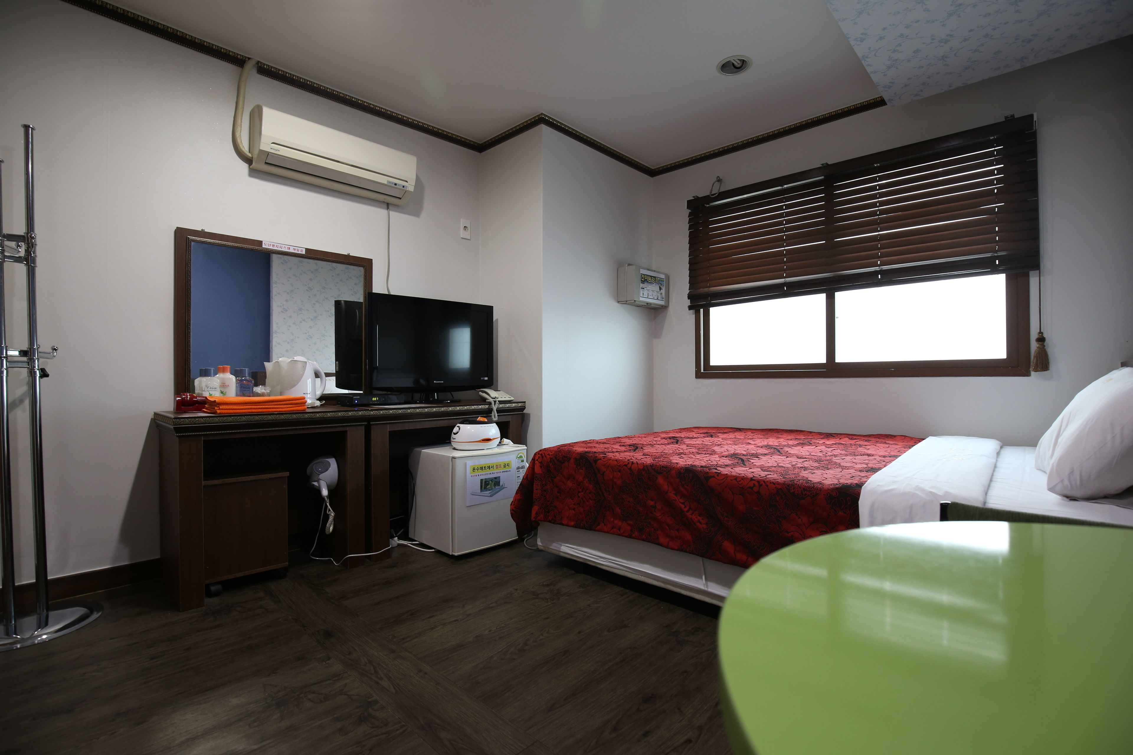 Une chambre d’hôtel avec un lit, un bureau avec une télévision, un climatiseur, une fenêtre avec des stores et un fauteuil vert.
