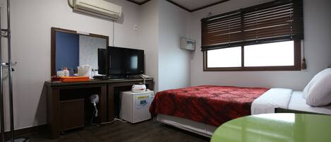 Une chambre d’hôtel avec un lit, un bureau avec une télévision, un climatiseur, une fenêtre avec des stores et un fauteuil vert.